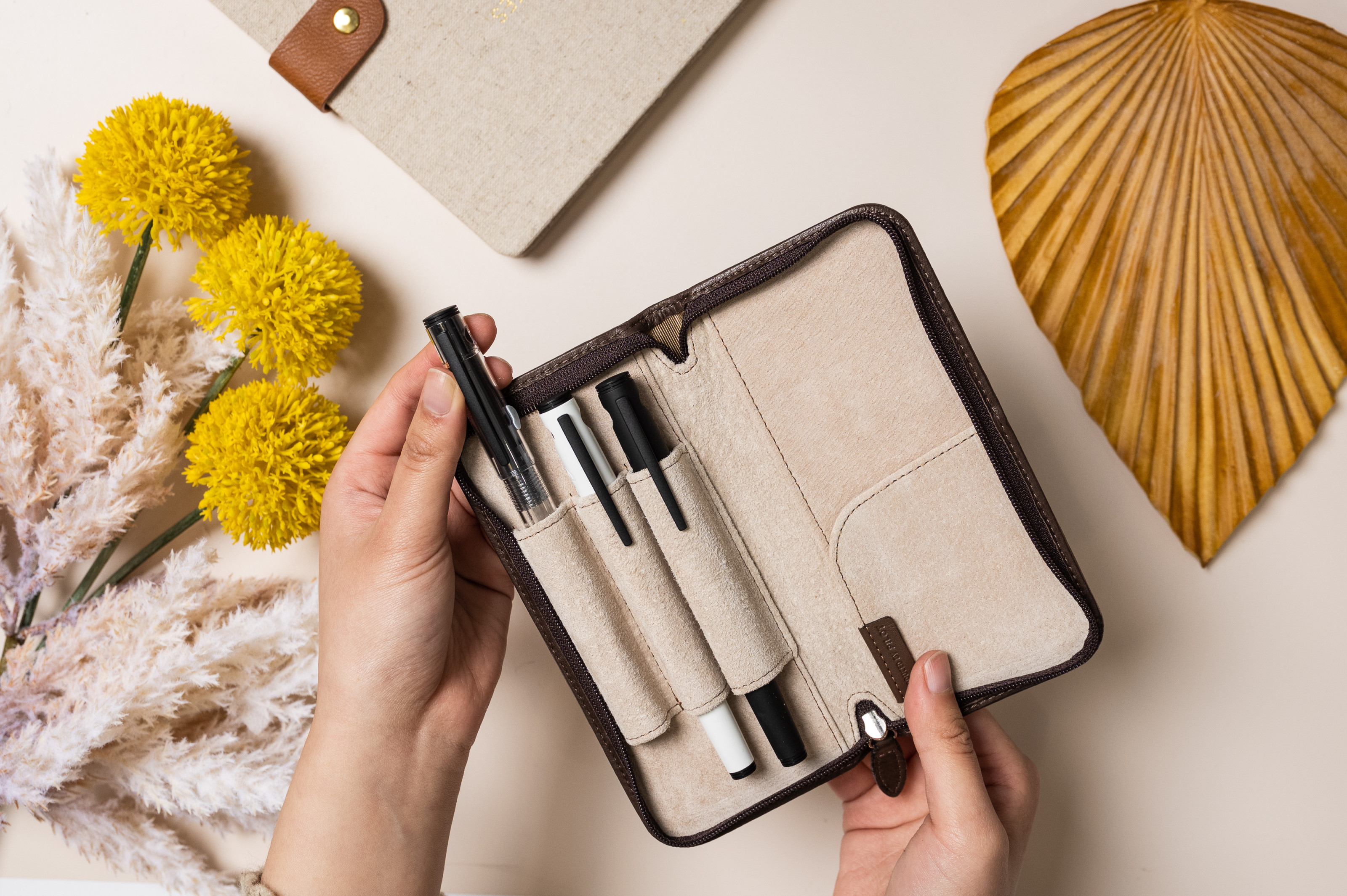 Three Pilot Explorer fountain pens in different colour barrels, a clear, white and black, sit in a Pilot Pensemble leather case, showcasing the collectable nature of fountain pens. A notebook with a linen cover can be seen in the top of the image. On the bottom right corner is a bottle of Pilot iroshizuku ink in deep pink, called kosumosu, which translates to cosmos in Japanese, a pink flower that looks similar to a daisy.