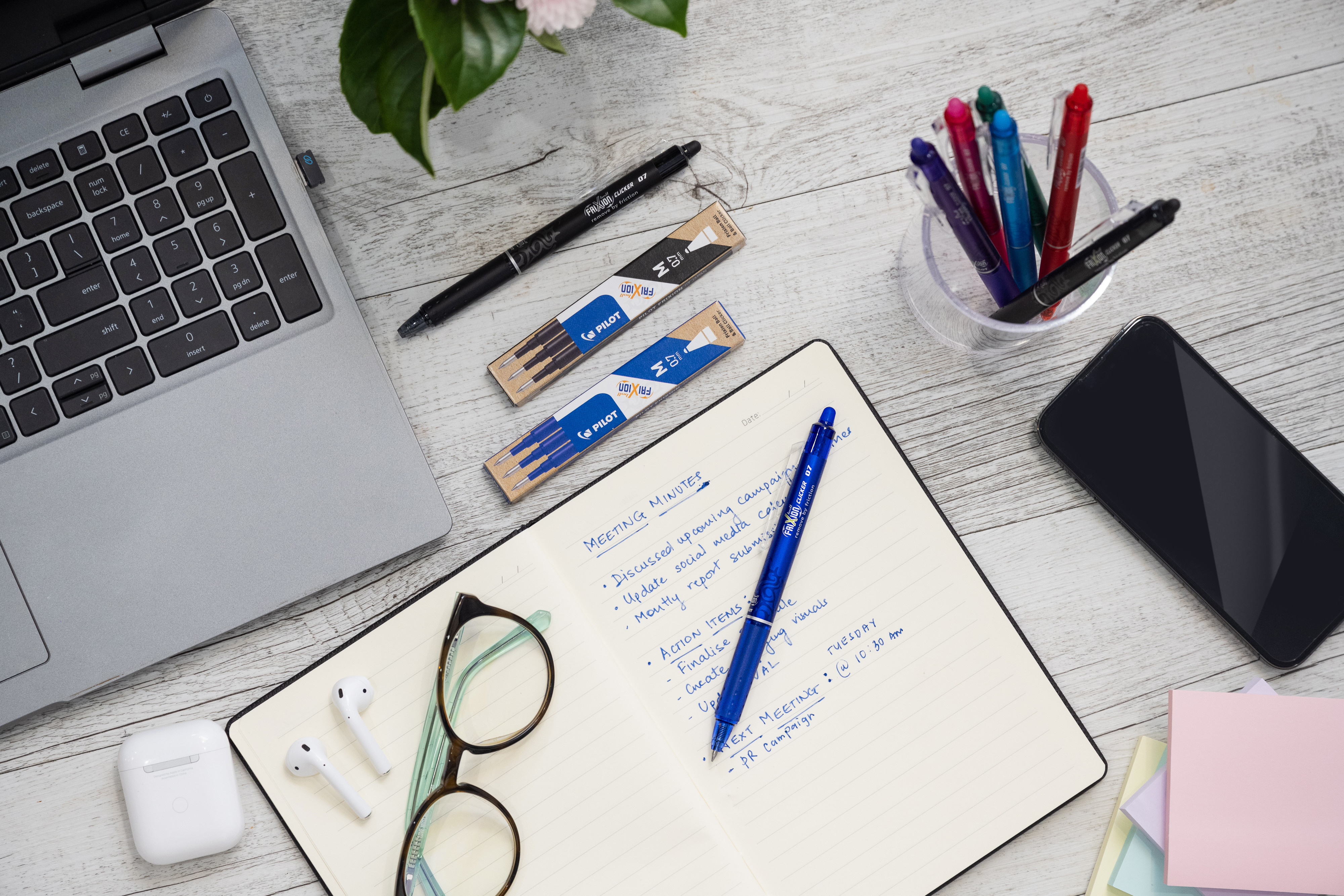 Laptop and notebook with blue FriXion Clicker Erasable Gel Pen on a desk with FriXion Erasable Gel Ink Refills pack in blue and black next to a laptop.