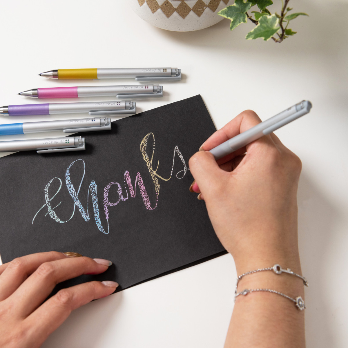 Close-up of a hand writing 'thanks' in cursive on black paper with a Juice Up Metallic Gel Pen, showcasing the pen's vibrant ink and precision for art.