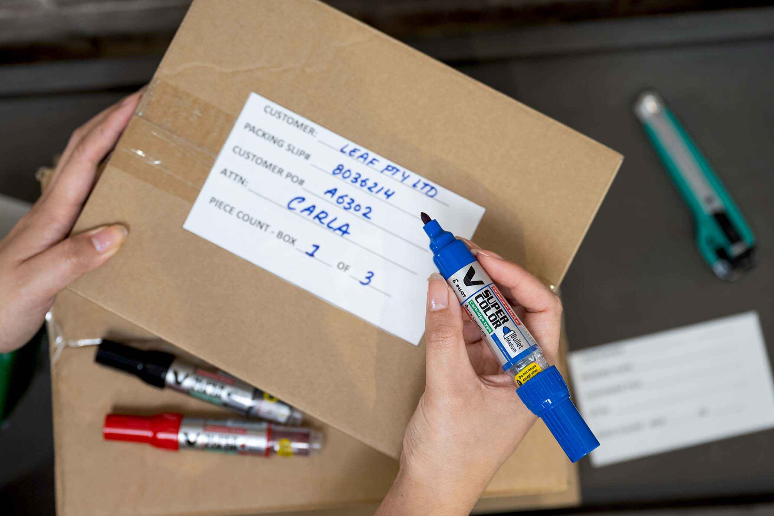 Address being written on a cardboard box with V Super Color Permanent Marker in blue.