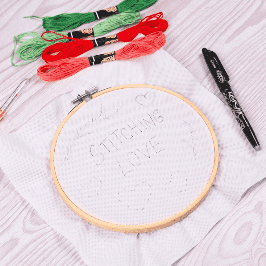 "Stitching Love" and designs drawn on fabric in a wooden embroidery hoop, written with a black FriXion Ball Gel pen.