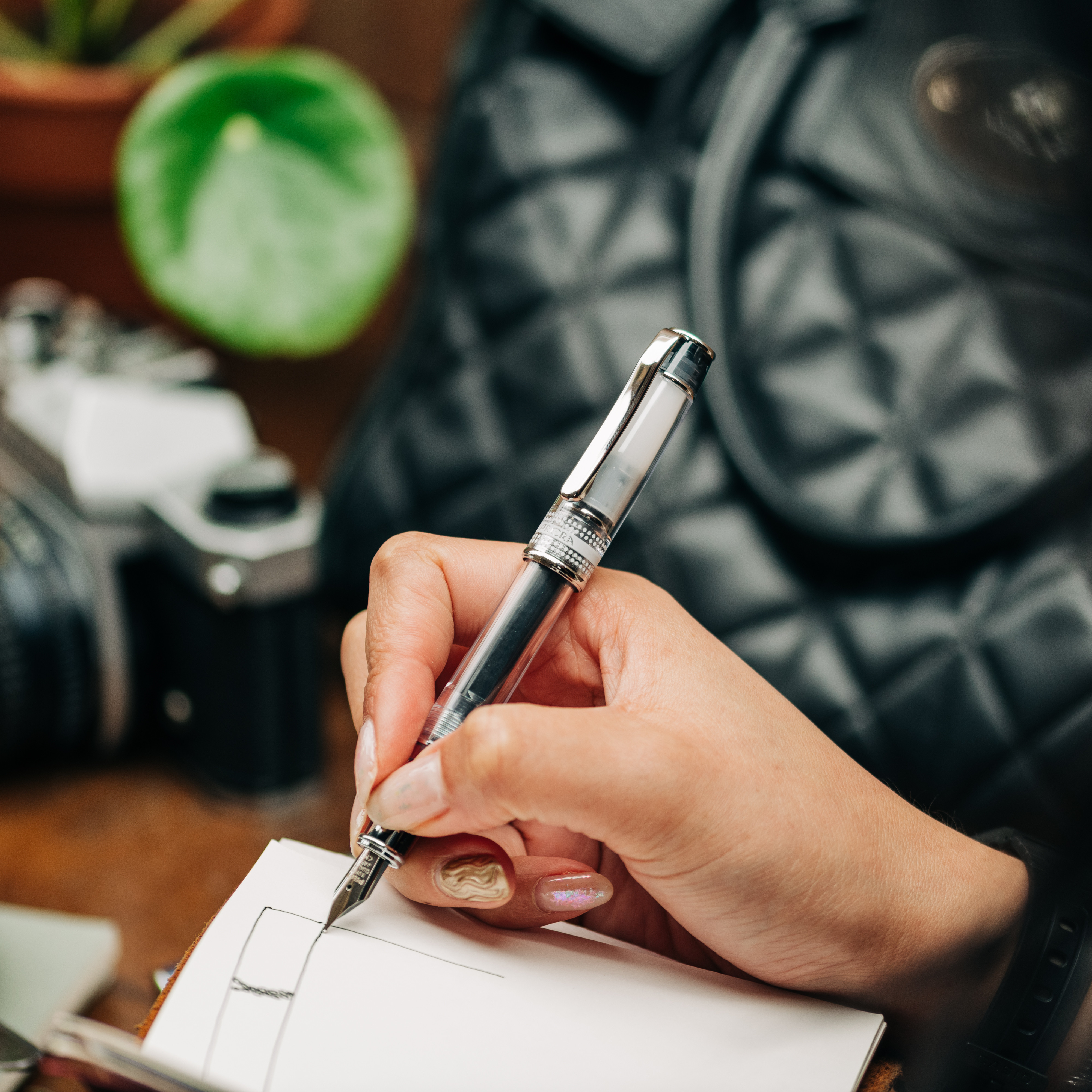 Pilot Prera Transparent fountain pen drawing in a notebook with black ink. A film camera, Pilea plant and leather bag can be seen in the background as the sketcher sits on a wooden table in a cafe.