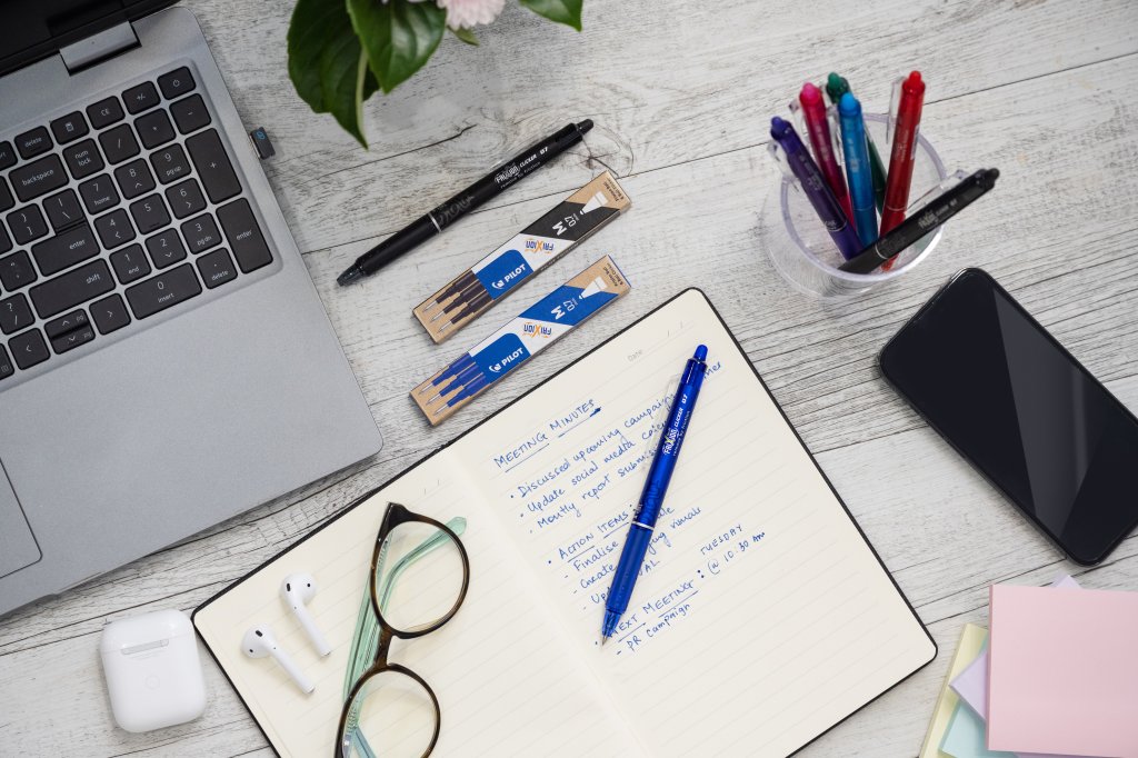 Pilot FriXion Clicker Erasable Pen in blue laying on a notepad on a desk with  FriXion Clicker in black and corresponding refills. More FriXion Clickers in many colours sit in a jar next to it.